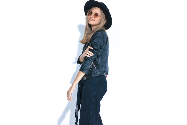 vista lateral de la joven sonriente sosteniendo el brazo en pose de moda y sonriendo, de pie aislado sobre fondo blanco en el estudio - Foto, Imagen