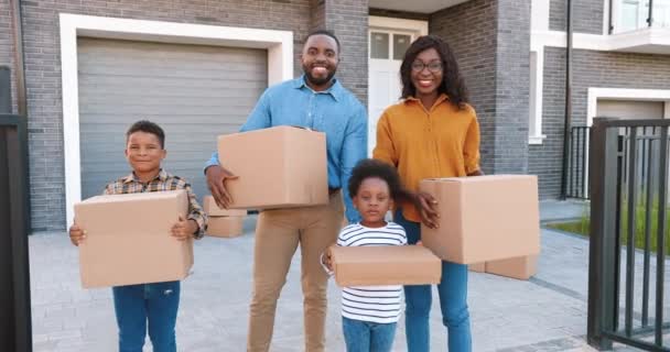 Porträt einer glücklichen afroamerikanischen Familie mit kleinen Kindern, die vor einem neuen Haus am Stadtrand steht und mit Kartons in der Hand lächelt. Fröhliche Eltern und Kinder beim Einzug in ein neues Zuhause. Siedlung am Stadtrand. - Filmmaterial, Video