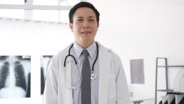 Fiatal ázsiai férfi orvos fehér orvosi egyenruhában, sztetoszkóppal nézi a kamerát, mosolyog és karok keresztbe, miközben videokonferencia hívás beteg az egészségügyi kórházban. Konzultációs és terápiás koncepció. - Felvétel, videó
