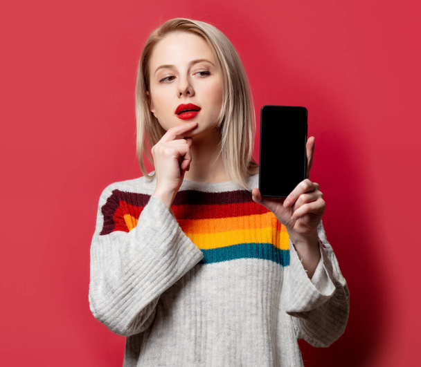 Belle blonde en pull avec téléphone portable sur fond rouge - Photo, image
