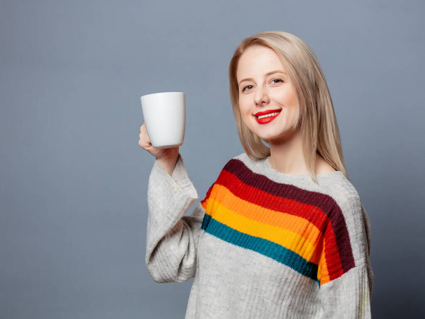 Krásná blondýna ve svetru s šálkem kávy na šedém pozadí - Fotografie, Obrázek