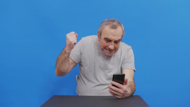 Happy isoisä voittaja mobiilisovellus urheilu veto tilalla älypuhelin ottaa positiivisia tunteita. Iloinen vanha mies nauraa juhlii voitto menestystä lue hyviä uutisia viesti. Eristetty sinisellä. - Materiaali, video
