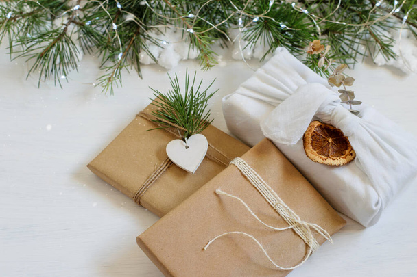 Cajas de regalo hechas a mano de Navidad envueltas en tela de reutilización y papel artesanal en estilo furoshiki japonés. Fondo blanco festivo con ramas de árbol de Navidad, espacio para copiar. Paquete de cuidado, conceptos bricolaje. - Foto, imagen