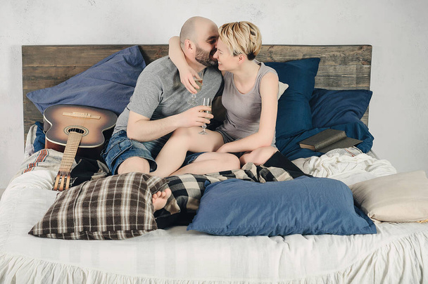 Un ragazzo con la barba e una ragazza bionda sono seduti sul letto. Chitarra, libro, champagne, emozioni, amore. Storia d'amore. - Foto, immagini