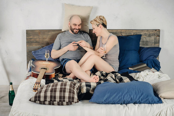 Facet z brodą i blondynką siedzą na łóżku. Gitara, książka, szampan, emocje, miłość. Historia miłości. - Zdjęcie, obraz