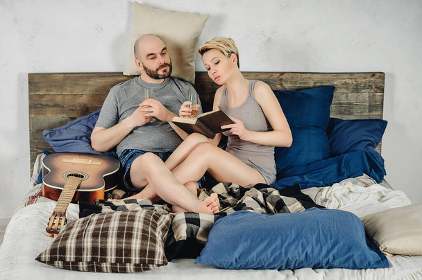 Egy szakállas fickó és egy szőke lány ül az ágyon. Gitár, könyv, pezsgő, érzelmek, szerelem. Szerelmi történet. - Fotó, kép
