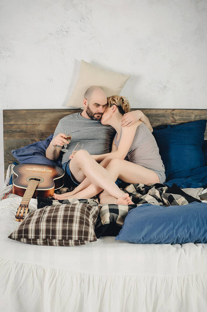 Een man met een baard en een blond meisje zitten op het bed. Gitaar, boek, champagne, emoties, liefde. Liefdesverhaal. - Foto, afbeelding
