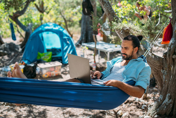 Человек работает на ноутбуке, когда лежит в гамаке. Молодой красавчик-фрилансер лежит в гамаке в палатке в горах среди деревьев и дистанционно работает в сети на ноутбуке. - Фото, изображение