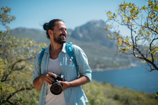 Мужчина турист, фотограф в горной роще с видом на море. Молодой красивый парень с туристическим рюкзаком и камерой на смотровой площадке в деревьях и смотрит на живописный вид на океан - Фото, изображение