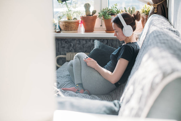 Дівчина сидить на дивані з телефоном у руках. Молода жінка розслабляється вдома, слухає музику на навушниках, дивиться відео на смартфон
 - Фото, зображення