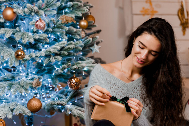 Дівчина відкриває різдвяний подарунок біля новорічної ялинки. Святкування Нового року. Сезонний продаж
 - Фото, зображення