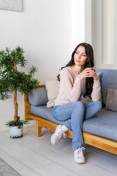 Νεαρή γυναίκα πίνει τσάι και ονειρεύεται να κάθεται στον καναπέ στο φωτεινό διαμέρισμα. - Φωτογραφία, εικόνα