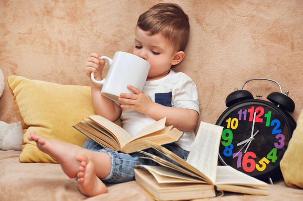 Un niño lindo en una camiseta blanca se sienta en un sofá en casa y bebe de una gran taza blanca cerca de un gran reloj despertador con números multicolores. - Foto, Imagen