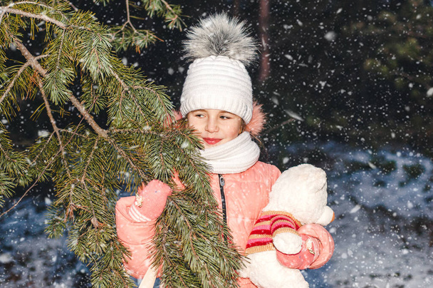 Grappig klein meisje in de winter besneeuwd Nieuwjaarsbos speelt met een speeltje witte beer. Winterportret van een kind. - Foto, afbeelding