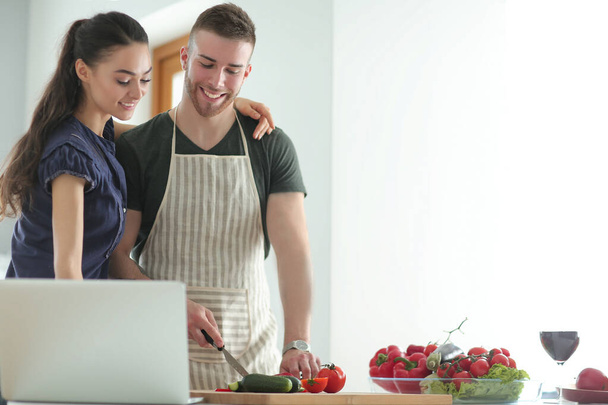 Young man cutting vegetables and woman standing with laptop in the kitchen - Φωτογραφία, εικόνα