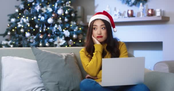 Mutsuz Asyalı, Noel Baba şapkalı güzel bir kadın dekore edilmiş modern evdeki koltukta oturuyor ve üzgün suratlı bir dizüstü bilgisayar kullanıyor. Noel arifesi kavramı. Yeni yıl kutlaması - Video, Çekim
