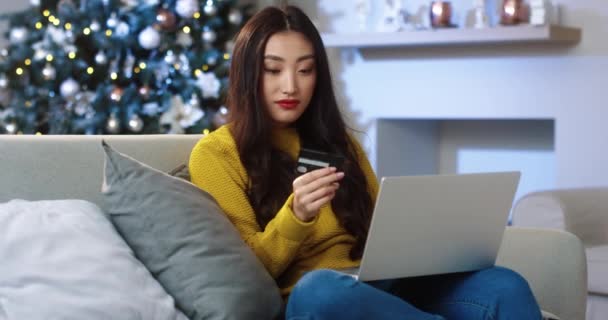 Happy excited Asian young woman sitting in decorated room on Christmas and buying Xmas presents online on laptop paying with credit card for purchase on internet. Праздничные. Концепция скидок - Кадры, видео