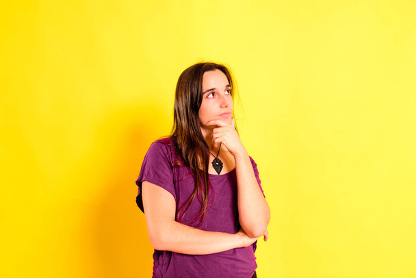 Mujer joven de pelo largo con expresión y gesto dudoso y pensativo, aislado sobre fondo amarillo. - Foto, imagen