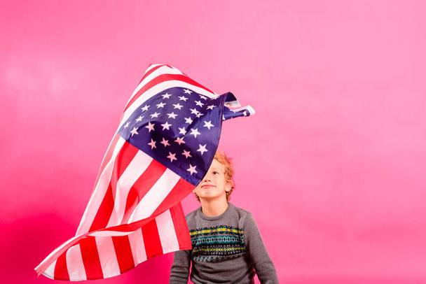 Blond boy under a floating American flag, isolated on a pink background. - Foto, imagen