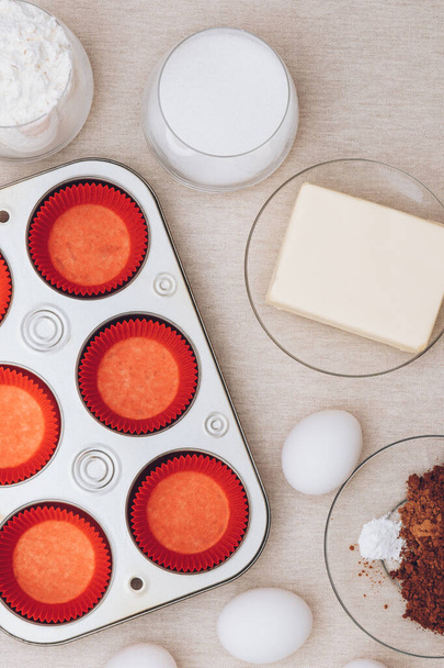 Rote Papierformen, Blech, Eier, ein Glas Mehl, ein Glas Milch, Kakao und ein Stück Butter auf der Tischdecke. Zutaten für Muffins. Ansicht von oben. - Foto, Bild