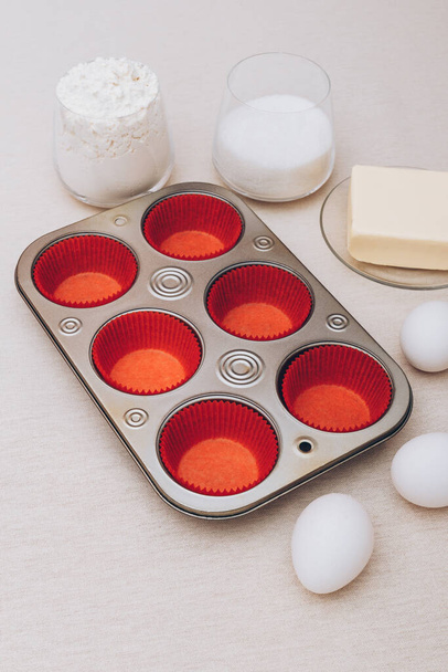 Red paper molds, metal baking tray, eggs, glass of flour, glass of milk and piece of butter on tablecloth.  Ingredients for cupcakes.  - Photo, Image