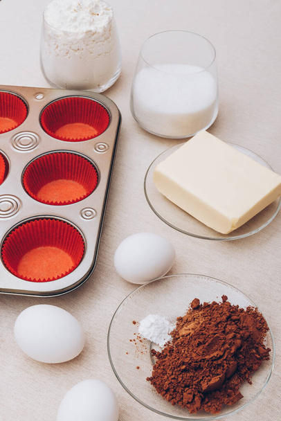 Red paper molds, metal baking tray, eggs, glass of flour, glass of milk, cacao and piece of butter on tablecloth. Ingredients for cupcakes.  - Photo, Image