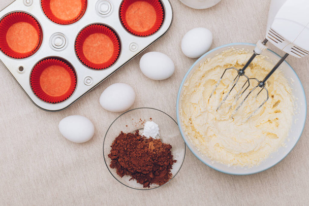 Βάζο για cupcakes σε ένα μπολ, κόκκινα καλούπια χαρτιού, μεταλλικό ταψί, αυγά, ένα ποτήρι αλεύρι, κακάο και μίξερ σε τραπεζομάντηλο. Προετοιμασία της διαδικασίας ψησίματος. - Φωτογραφία, εικόνα