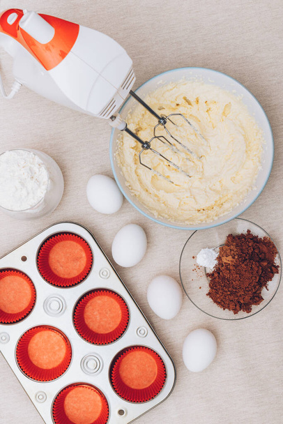 Dough for cupcakes in a bowl, red paper molds, metal baking tray, eggs, glass of flour, cacao and mixer on tablecloth. Preparing process to baking. - Photo, Image