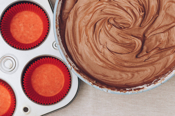 Kakao hamuru kap kekler, kırmızı kağıt kalıplar ve masa örtüsünün üzerindeki metal pişirme tepsisi için. Pişirme işlemi hazırlanıyor. Üst görünüm. - Fotoğraf, Görsel