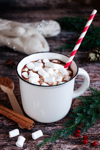 White cup of hot spiced cacao or chocolate with marshmallows and a striped paper straw on a rustic wooden tabletop with fir branches and knitted gloves in the background.  - Foto, Imagem