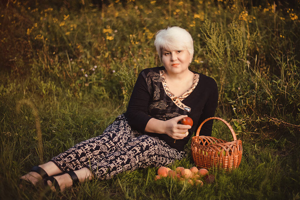 Egy szőke nő ül egy kosár friss körtével és őszibarackkal az erdő zöld gyepén.. - Fotó, kép