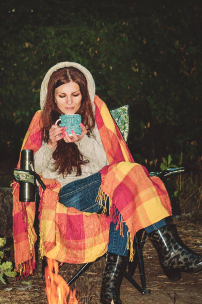 Een prachtig roodharig meisje in de avond herfst bos zit in een stoel en een ruitje rond een vuur en drinkt thee. - Foto, afbeelding