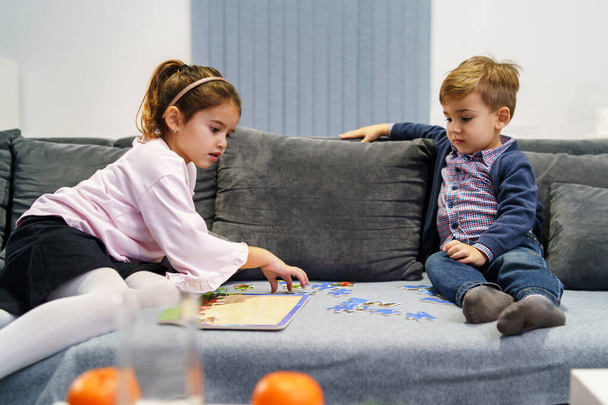 Двое детей мальчик и девочка три брата и сестра четырех или пяти лет играют дома на диване-кровати с головоломками, изучая развивающую игру - Фото, изображение