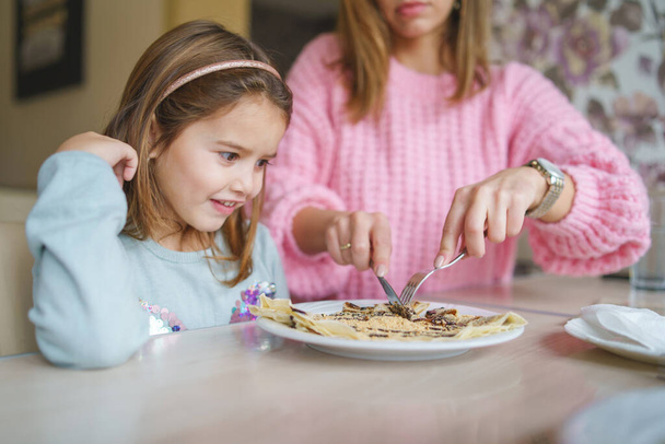 Roztomilé holčička sedí u stolu, zatímco její matka krájí sladké palačinky se smetanou dcera jíst - Fotografie, Obrázek