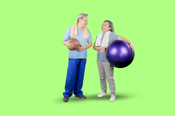 Full length of elderly couple wearing sportswear while holding pilates ball and basketball and standing together in the studio with green screen background - Foto, afbeelding