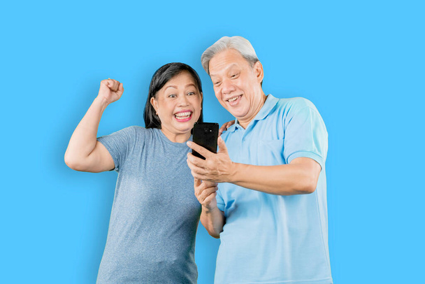 Happy old couple reading good news on cellphone while standing in the studio with blue background - Fotoğraf, Görsel
