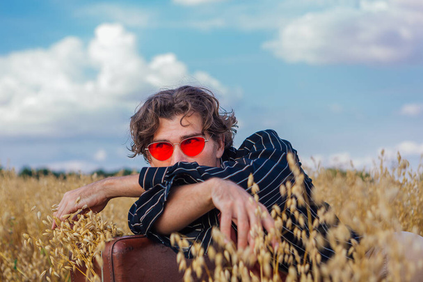 Rural Countryside Scene. Tall handsome man dressed in a black shirt and pink sunglasses laying at golden oat field with brown vintage leather suitcase full of ears of oats. Summer landscape with blue sky - Fotografie, Obrázek