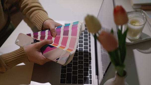 Captura recortada de manos de diseñador sosteniendo la muestra de color mientras trabaja con el ordenador portátil en la oficina en casa  - Foto, Imagen