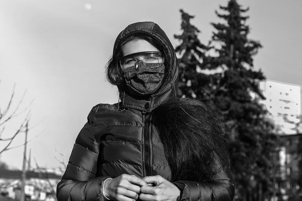 Una mujer en la primavera en la calle con una máscara médica verde casera y gafas que protegen las membranas mucosas del ojo durante la cuarentena debido al coronavirus. 2019-nCoV Foto en blanco y negro. - Foto, Imagen
