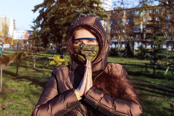 Una mujer en la primavera en la calle con una máscara médica verde casera y gafas que protegen las membranas mucosas del ojo durante la cuarentena debido al coronavirus. 2019-nCoV - Foto, Imagen