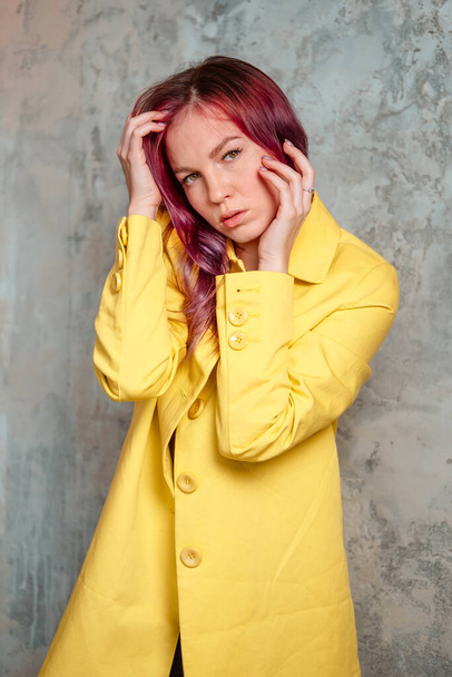 Beautiful woman with pink hair dressed in yellow raincoat touching her face and posing indoor near the concrete wall. - Foto, afbeelding