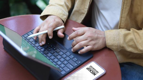 Vista de cerca de manos masculinas tecleando el teclado de la tableta de la pluma en la mesa de centro con teléfono inteligente en el jardín - Foto, Imagen
