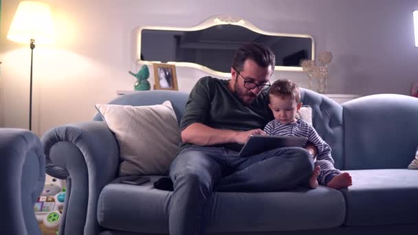 Padre y lindo bebé sentado en el sofá en casa jugando juntos en la tableta digital - Metraje, vídeo