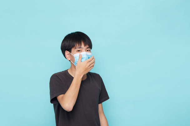 Primo piano del giovane asiatico che indossa una maschera facciale contro la covid-19 su sfondo blu isolato, concetto coronavirus - Foto, immagini
