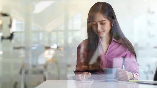 Blick durch Glaswand: Geschäftsfrau entspannt mit Smartphone und Kaffeetasse - Foto, Bild