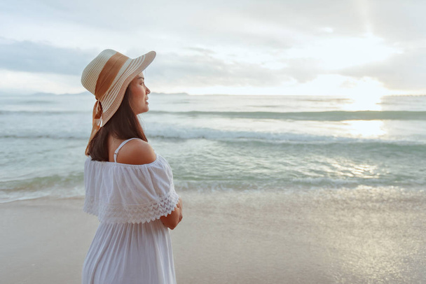 Azjatka ubrana w stylowy kapelusz i ubrania patrząc na ocean i ciesząc się pięknym zachodem słońca na plaży. Wakacje letnie i koncepcja podróży. - Zdjęcie, obraz