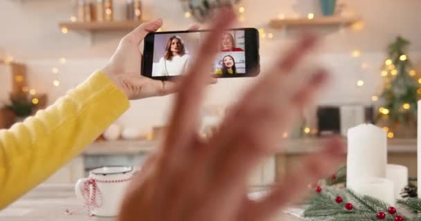 Female hand hold smartphone with online video call talking with multi-ethnic Asian and Caucasian friends celebrating New Year in decorated kitchen. Winter holidays. Close up. multiple chat - Footage, Video