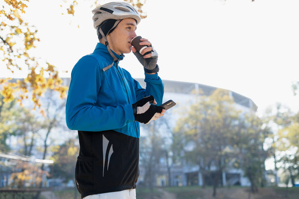 A young man in a bicycle helmet drinks hot coffee from a glass and holds a smartphone in his hands. Food delivery around the city on bicycles. - Valokuva, kuva