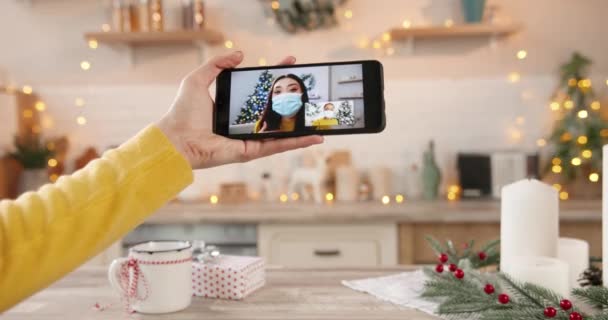 Close up of woman hand holds smartphone with multiple video conference with mixed-races female friends in medical masks at decorted home in quarantine. New Years Eve. Holiday season concept - Footage, Video