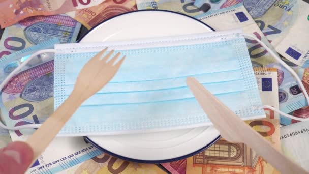 Coronavirus lockdown concept. Cutting a medical mask with a wooden knife and fork on a white plate with a blue border. Euro currency banknotes background. Close up - Video